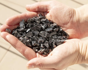May include: A handful of black, irregularly shaped shungite stones. The stones are held in cupped hands, with the text "Shungite Zazhogino" visible. The stones have a reflective, dark appearance.