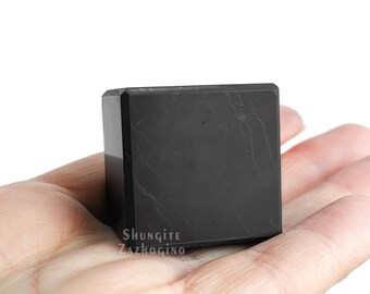 May include: A black, cube-shaped piece of Shungite, a type of metamorphic rock, with a smooth, polished surface. The stone is resting on a hand.