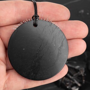 Puede incluir: Un colgante redondo de shungita negro con un cord&oacute;n negro. El colgante tiene una superficie lisa y pulida con estr&iacute;as visibles. El colgante est&aacute; siendo sostenido en una mano.