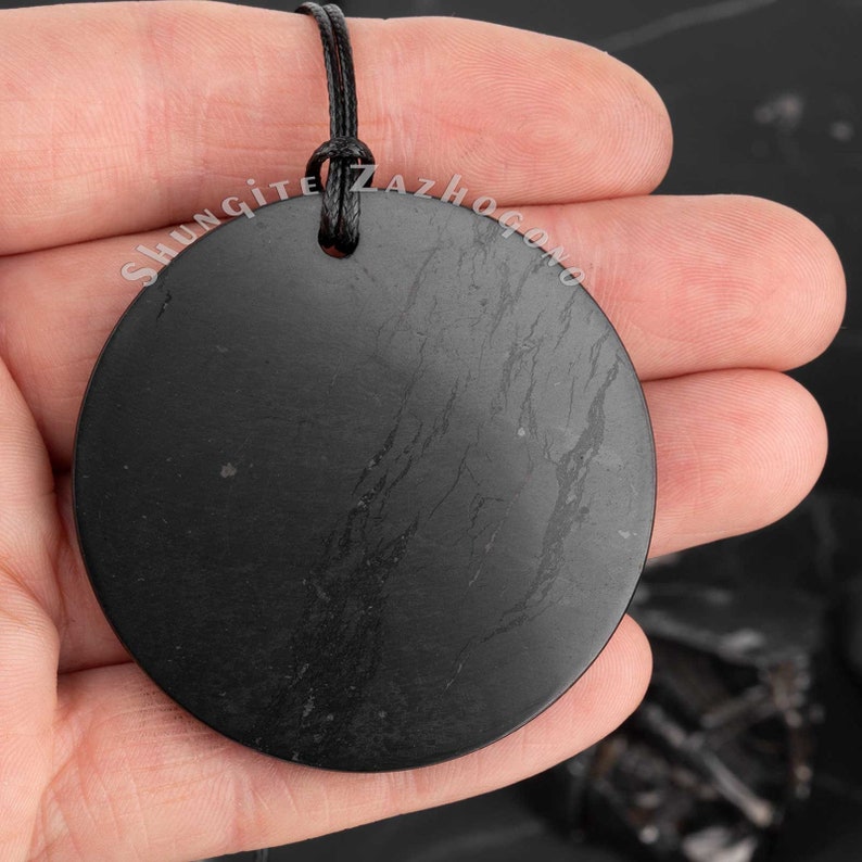 Puede incluir: Un colgante redondo de shungita negro con un cord&oacute;n negro. El colgante tiene una superficie lisa y pulida con estr&iacute;as visibles. El colgante est&aacute; siendo sostenido en una mano.