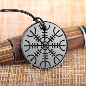 May include: A black shungite pendant with a silver Vegvisir symbol, a Norse directional symbol, engraved on it. The pendant is on a black cord and is resting on a woven surface. The text "Shungite Zazhogino" is visible on the pendant.