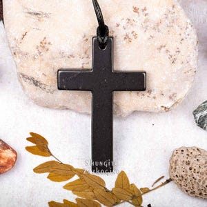May include: Black shungite cross pendant on a black cord. The cross is on a white background with small rocks and dried leaves.
