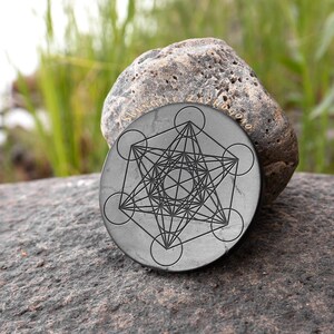 Puede incluir: Un posavasos redondo de piedra negra con un diseño geométrico grabado en su superficie. El diseño presenta una estrella con múltiples círculos superpuestos. El posavasos está apoyado sobre una roca gris con las palabras "Shungite Zahorgon" inscritas en la roca.