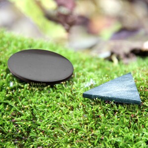 以下が含まれることがあります： 緑の苔の上に、黒と緑の磨かれた石が2つ置かれています。大きな石は丸く、小さな石は三角形です。苔の上に「Shungite Zakologing」という文字が見えます。