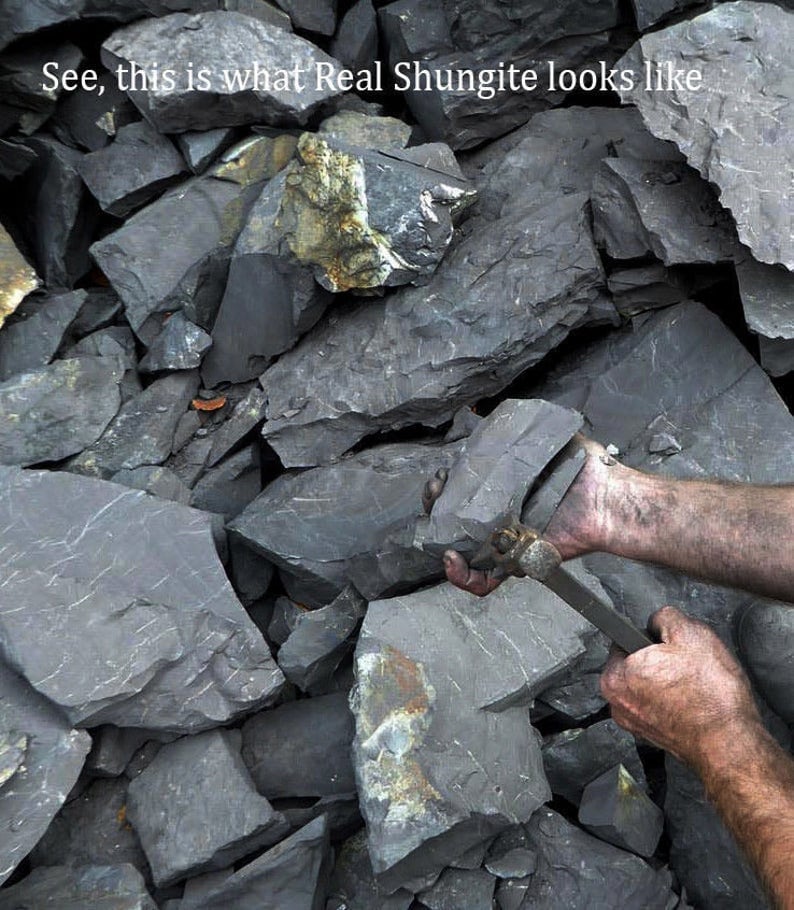 Puede incluir: Primer plano de una pila de rocas gris oscuro. Unas manos sostienen una roca y una herramienta, probablemente para partir la roca. El texto "See, this is what Real Shungite looks like" es visible en la parte superior de la imagen.