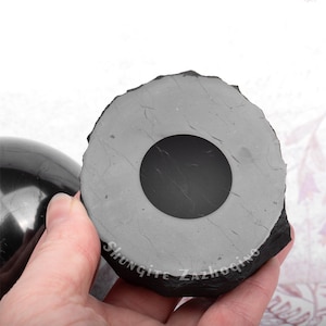 Puede incluir: Una esfera de shungita negra con un agujero en el centro, descansando sobre una base de shungite gris. La base tiene la palabra "Shungite" inscrita en ella.