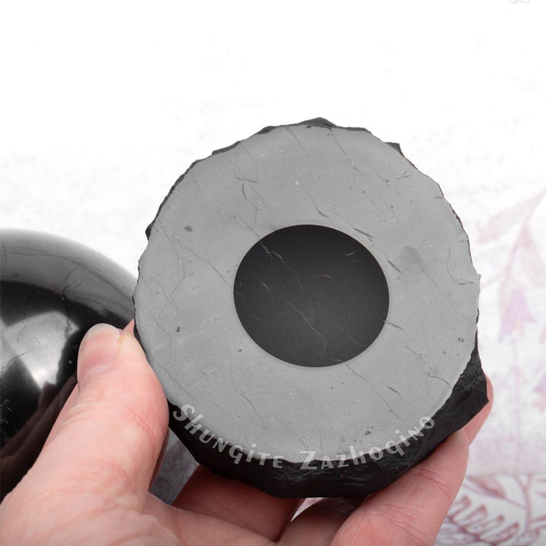 Puede incluir: Una esfera de shungita negra con un agujero en el centro, descansando sobre una base de shungite gris. La base tiene la palabra "Shungite" inscrita en ella.