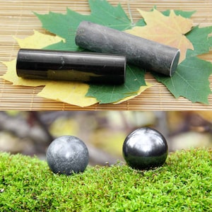 May include: Two black and gray smooth stone spheres and two cylindrical stones, one black and one gray, on a green mossy surface and a brown woven surface with yellow and green leaves.