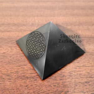 May include: Black Shungite pyramid with a carved flower of life design. The pyramid is on a wooden surface. The text "Shungite Zazhogino" is visible on the pyramid.