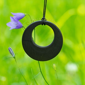 May include: A black shungite pendant with a round, ring-shaped design hangs from a black cord against a green background. The pendant is surrounded by purple wildflowers.