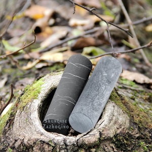 Può includere: Due pietre nere lucidate con venature bianche, una è liscia e l'altra ha una texture più ruvida. Le pietre sono appoggiate su un ceppo d'albero ricoperto di muschio. Il testo "Shungite Zazhogino" è visibile in basso a sinistra dell'immagine.