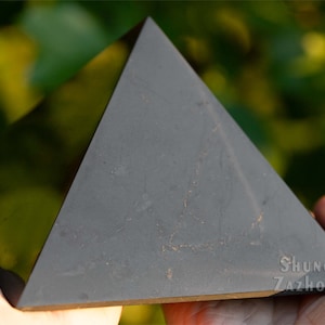 May include: A black pyramid-shaped piece of Shungite, a type of carbon-rich metamorphic rock, with the text "Shungite Zazhogino" on the bottom.