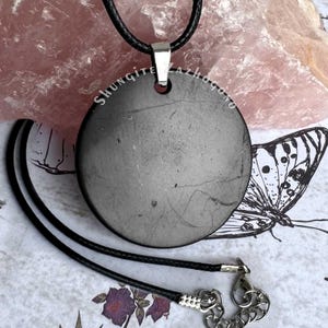 Puede incluir: Un colgante redondo de shungita negra en un collar de cord&oacute;n negro. El colgante mide aproximadamente 5 cm de di&aacute;metro y tiene un aro de plata. El collar mide unos 45 cm de largo y tiene un cierre de plata. El colgante est&aacute; sobre un fondo blanco con un cristal de cuarzo rosa y una ilustraci&oacute;n de mariposa.