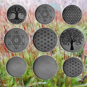 Puede incluir: Nueve posavasos redondos de pizarra gris con varios diseños grabados en negro. Los diseños incluyen un árbol de la vida, una flor de la vida y un patrón geométrico. Los posavasos están dispuestos en círculo sobre un lecho de musgo verde.
