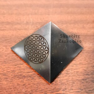 May include: A black pyramid made of Shungite with a flower of life design engraved on one side. The pyramid is labeled "Shungite Zazhogino".
