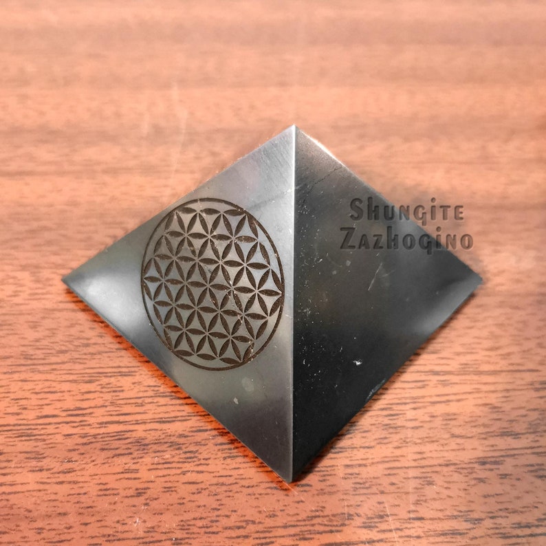 May include: A black pyramid made of Shungite with a flower of life design engraved on one side. The pyramid is labeled "Shungite Zazhogino".