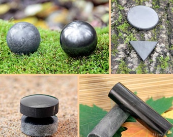 May include: A collection of polished black shungite stones in various shapes, including a sphere, a disc, a pyramid, and a cylinder. The stones are displayed on different surfaces, including moss, wood, and sand.