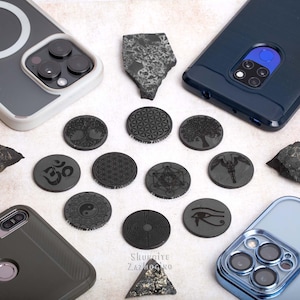 May include: A collection of 12 black round phone grips with various engraved designs, including a tree, a flower of life, an Om symbol, a yin yang, a labyrinth, an eye, and a bird. The grips are arranged on a light gray surface with two black rocks and two cell phones.