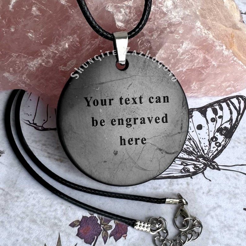 Puede incluir: Un collar con un colgante redondo negro en un cord&oacute;n negro. El colgante est&aacute; hecho de shungita y tiene el texto "Your text can be engraved here" grabado.