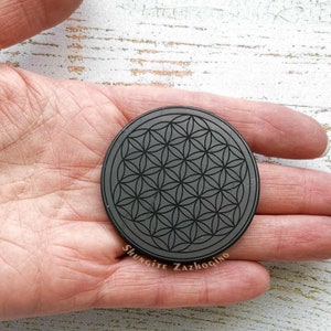 May include: A black shungite stone with a flower of life design engraved on it. The stone is round and about 2 inches in diameter. The text "Shungite Zazhogino" is visible on the stone.