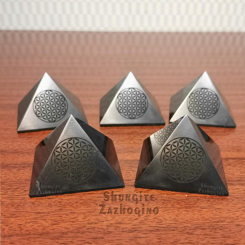 May include: Five black shungite pyramids with a flower of life design engraved on each side. The pyramids are on a wooden surface. The text "Shungite Zazhogino" is visible on the bottom of two of the pyramids.