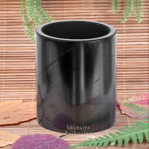 Pode incluir: Recipiente cilíndrico de pedra shungite preta. O recipiente é liso e tem uma aparência natural, marmorizada. Está sentado em um tapete de bambu tecido com folhas de outono.