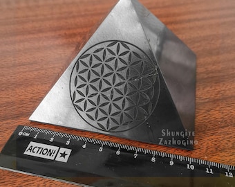 May include: Black shungite pyramid with a flower of life design engraved on one side. The pyramid is approximately 7 cm wide and is sitting on a wooden surface. The text "Shungite Zazhogino" is printed on the pyramid.