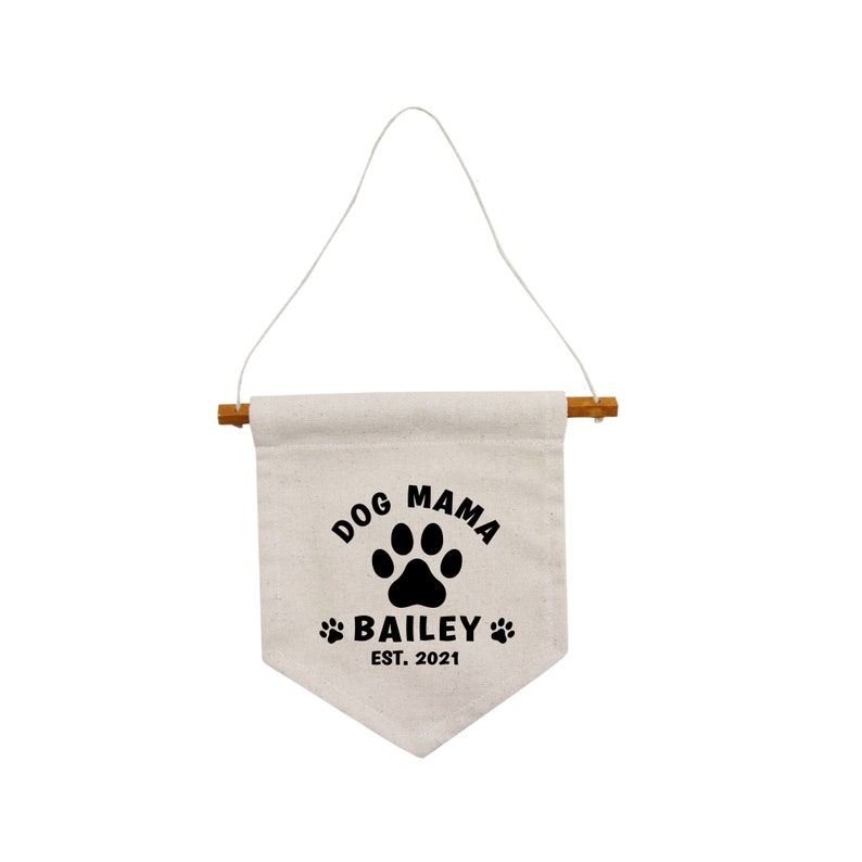 May include: A white canvas banner with a black paw print and the text "Dog Mama Bailey Est. 2021". The banner is hanging from a wooden dowel rod with a white string.
