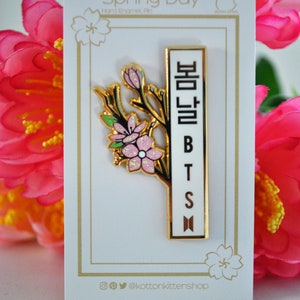 May include: A gold-colored enamel pin with a floral design and the text "Spring Day" and "BTS". The pin features a branch with pink flowers and a white rectangular section with Korean characters and the letters "BTS".