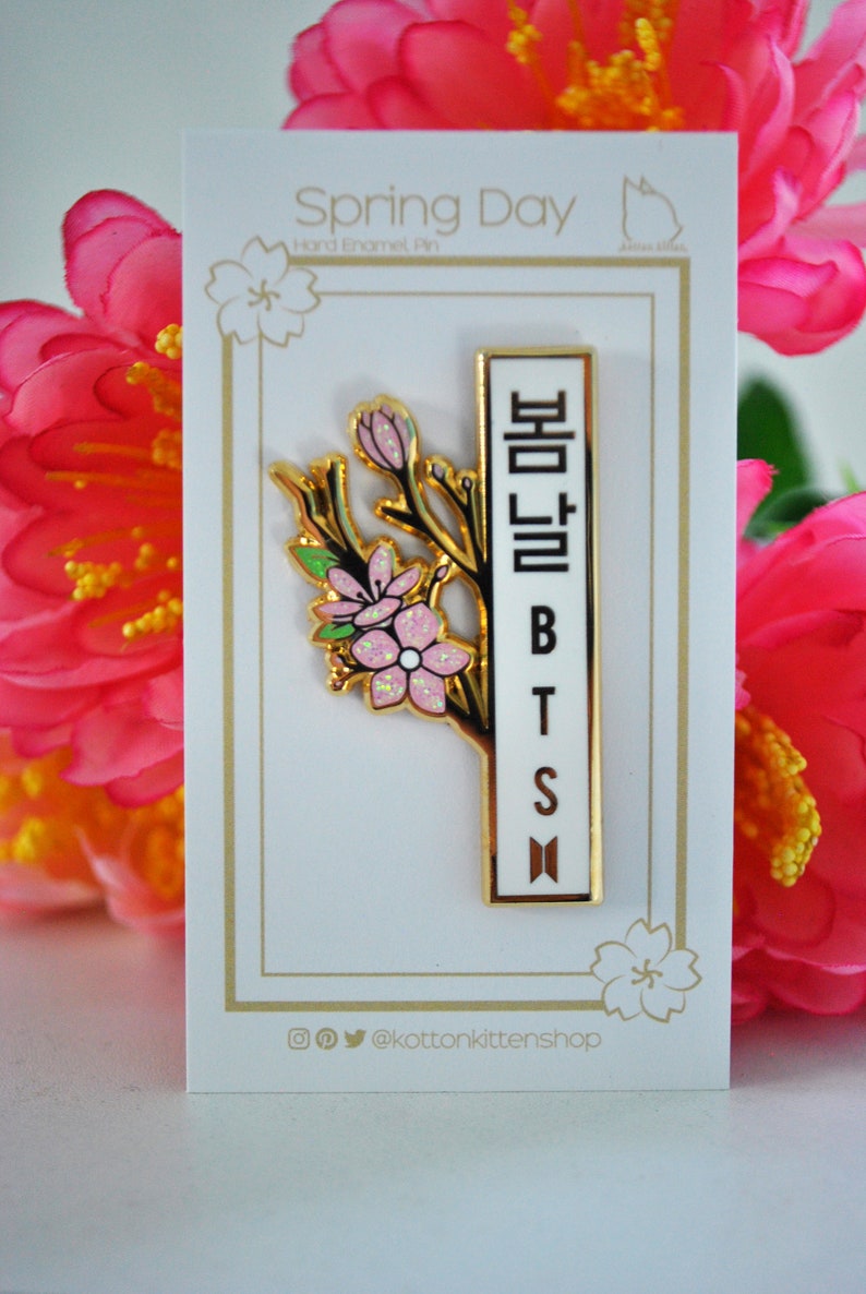 May include: A gold-colored enamel pin with a floral design and the text "Spring Day" and "BTS". The pin features a branch with pink flowers and a white rectangular section with Korean characters and the letters "BTS".