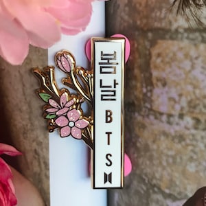 May include: A gold-colored pin featuring a floral design with pink glitter accents and the text "BTS" in black. The pin also includes Korean text and a small logo. The pin is set against a white background.