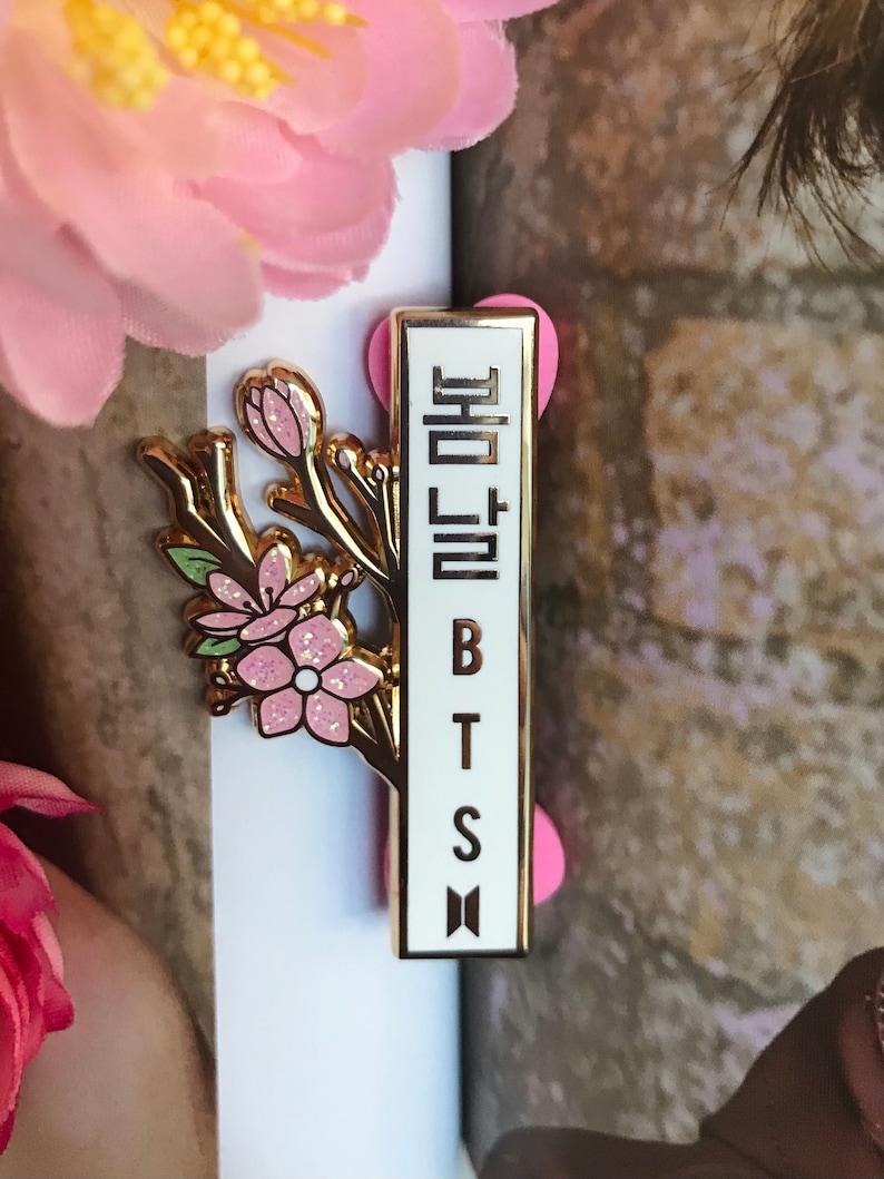 May include: A gold-colored pin featuring a floral design with pink glitter accents and the text "BTS" in black. The pin also includes Korean text and a small logo. The pin is set against a white background.