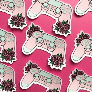 May include: Pink and white Playstation controller with a floral design. The controller has a pink background with white buttons and a white border. The floral design is in shades of pink and red.