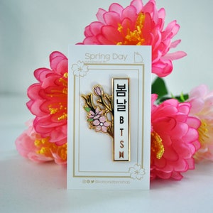May include: A gold-colored enamel pin with a floral design and the text "봄날 BTS" in Korean. The pin is on a white card with the words "Spring Day" and surrounded by pink flowers.