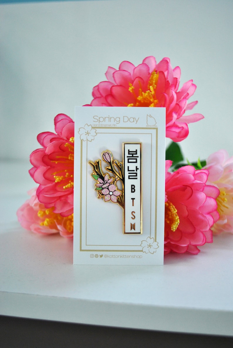 May include: A gold-colored enamel pin with a floral design and the text "봄날 BTS" in Korean. The pin is on a white card with the words "Spring Day" and surrounded by pink flowers.
