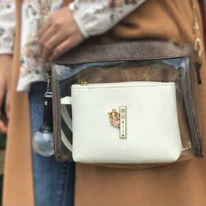 May include: A white zippered pouch with a gold-colored zipper and a floral pin. The pin has pink and green accents and the text "BTSN" in a vertical gold bar. The pouch is inside a clear bag with a brown top.
