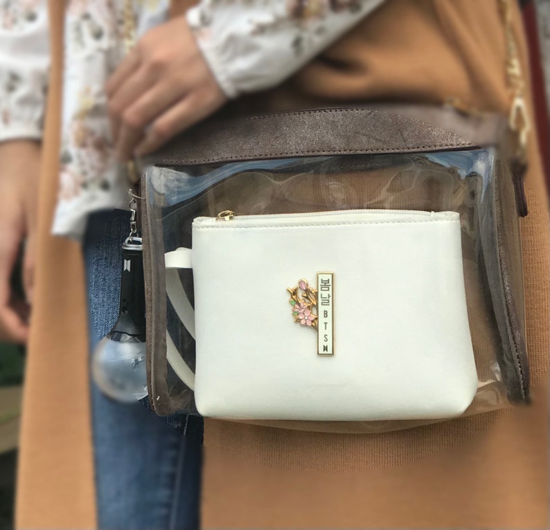 May include: A white zippered pouch with a gold-colored zipper and a floral pin. The pin has pink and green accents and the text "BTSN" in a vertical gold bar. The pouch is inside a clear bag with a brown top.