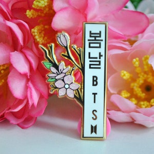 May include: A rectangular pin with a gold border and the text "봄날 BTS" in black. The pin features a branch with pink flowers and green leaves. The background is a cluster of pink flowers.