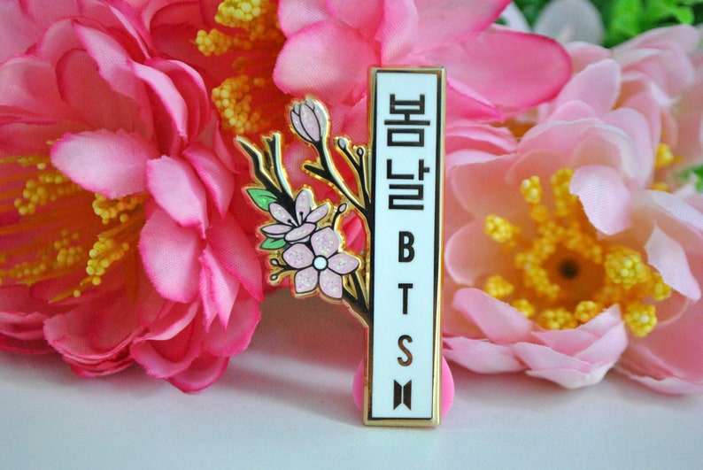 May include: A rectangular pin with a gold border and the text "봄날 BTS" in black. The pin features a branch with pink flowers and green leaves. The background is a cluster of pink flowers.