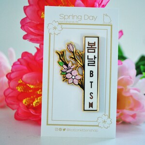 May include: A gold-colored enamel pin featuring a floral design with pink blossoms and green leaves. The pin has a white vertical bar with the text "BTS" and Korean characters. The pin is on a white card with the text "Spring Day".