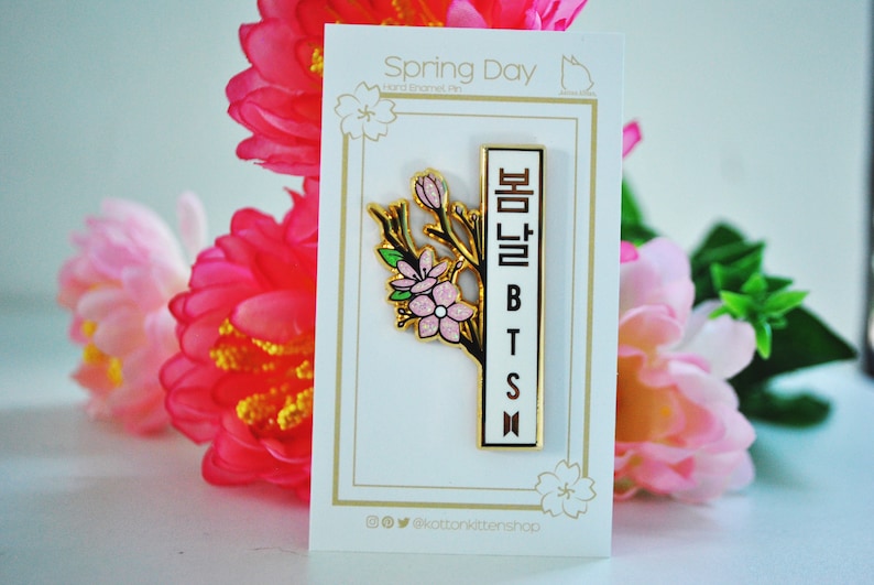 May include: A gold-colored enamel pin featuring a floral design with pink blossoms and green leaves. The pin has a white vertical bar with the text "BTS" and Korean characters. The pin is on a white card with the text "Spring Day".