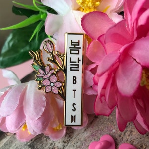 May include: A gold-colored pin with a floral design and the Korean text "봄날" and the letters "BTS" in white. The pin is surrounded by pink flowers and green leaves. Two pink heart-shaped pin backs are visible.