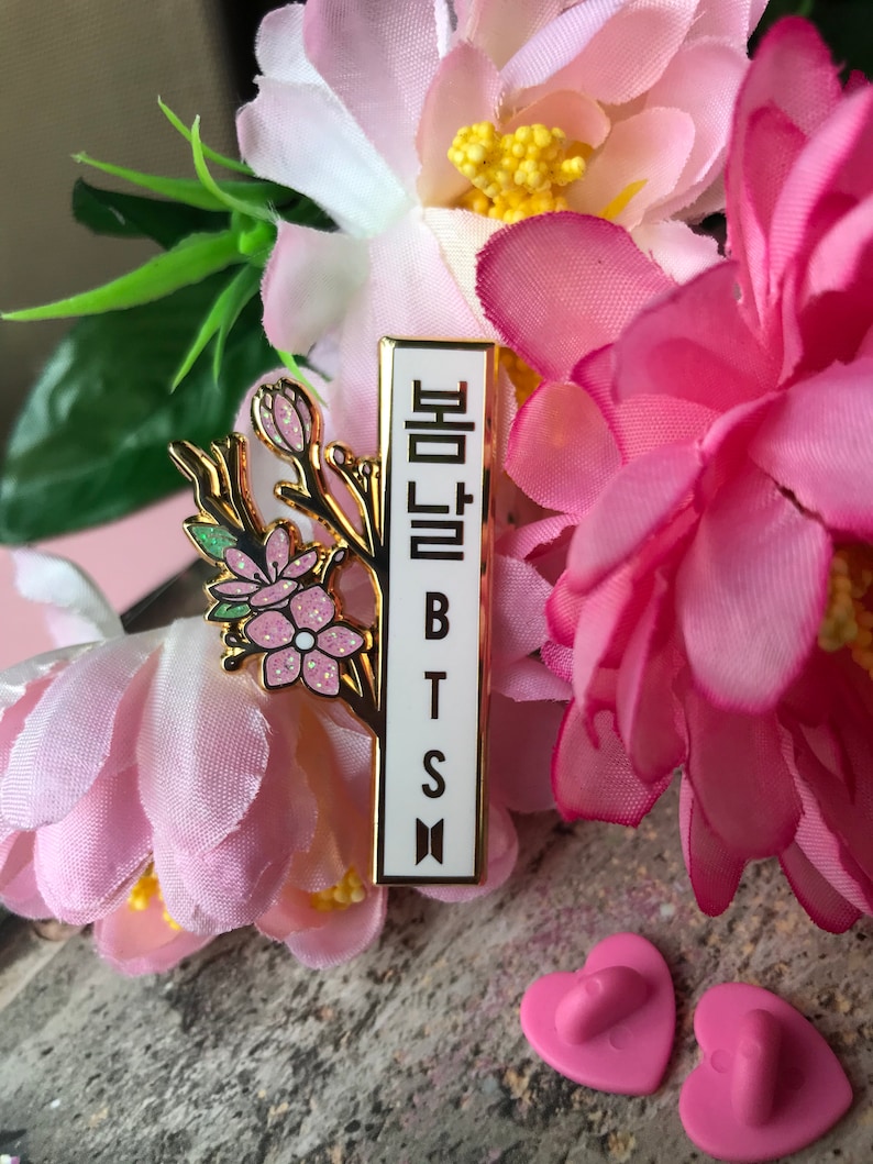 May include: A gold-colored pin with a floral design and the Korean text "봄날" and the letters "BTS" in white. The pin is surrounded by pink flowers and green leaves. Two pink heart-shaped pin backs are visible.
