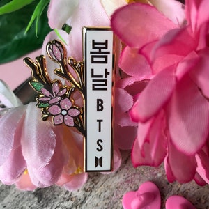 May include: A rectangular enamel pin with the text "봄날 BTS" in Korean and English. The pin features a gold border and a floral design with pink and white flowers. The pin is surrounded by pink flowers.