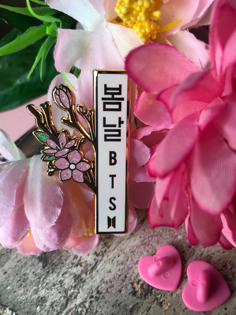 May include: A rectangular enamel pin with the text "봄날 BTS" in Korean and English. The pin features a gold border and a floral design with pink and white flowers. The pin is surrounded by pink flowers.
