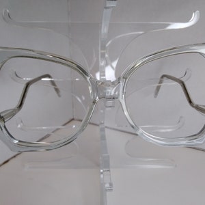May include: Clear plastic framed glasses with a geometric shape. The glasses are on a white display stand.