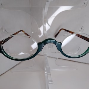 May include: A pair of round, green-framed eyeglasses with clear lenses. The frames are made of a dark green plastic and have a tortoise shell pattern on the arms.