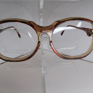 May include: A pair of brown and tortoiseshell eyeglasses with round lenses. The glasses are on a clear plastic stand.