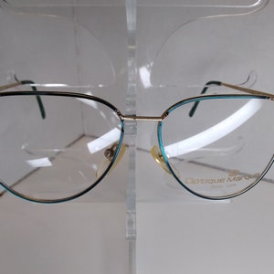 May include: Gold-tone cat-eye eyeglasses with a teal trim. The glasses have clear lenses and the brand name "Optique Marquis" is printed on the right lens.