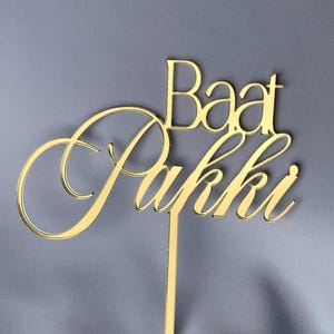 Könnte beinhalten: Goldfarbener Tortendekor mit den Worten "Baat Pakki" in einer kursiven Schrift.